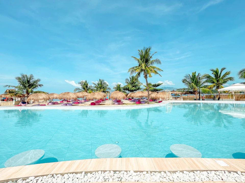 A pool with clear blue water surrounded by palm trees and straw umbrellas under a sunny sky.