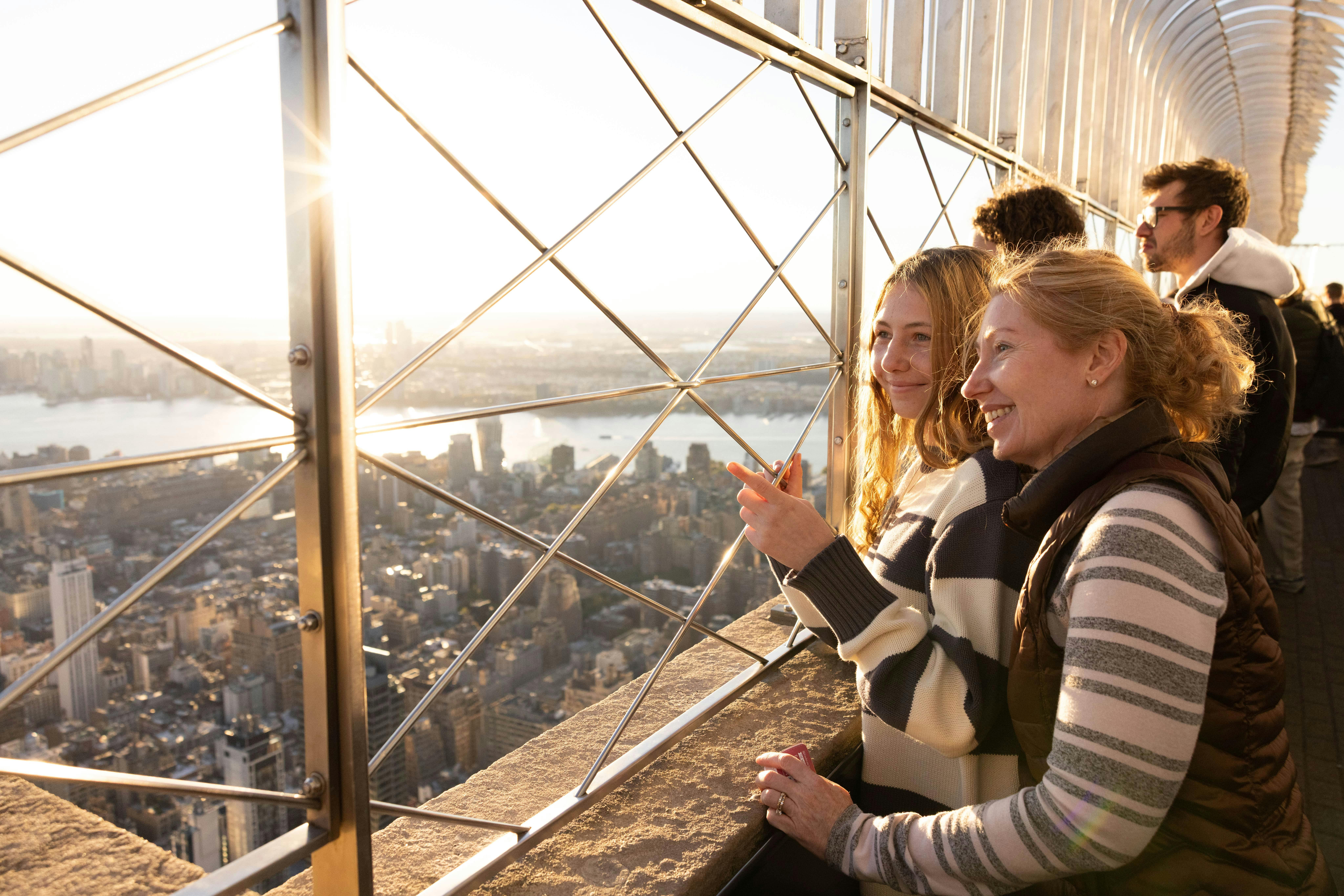 Two people smile and look out from an observation deck with a cityscape view through a metal fence, at sunset.