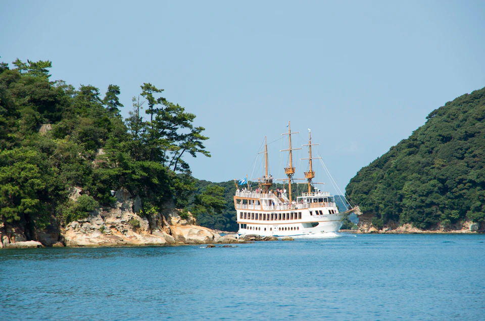 A white ship with sails navigates near a forested coastline under a clear blue sky.