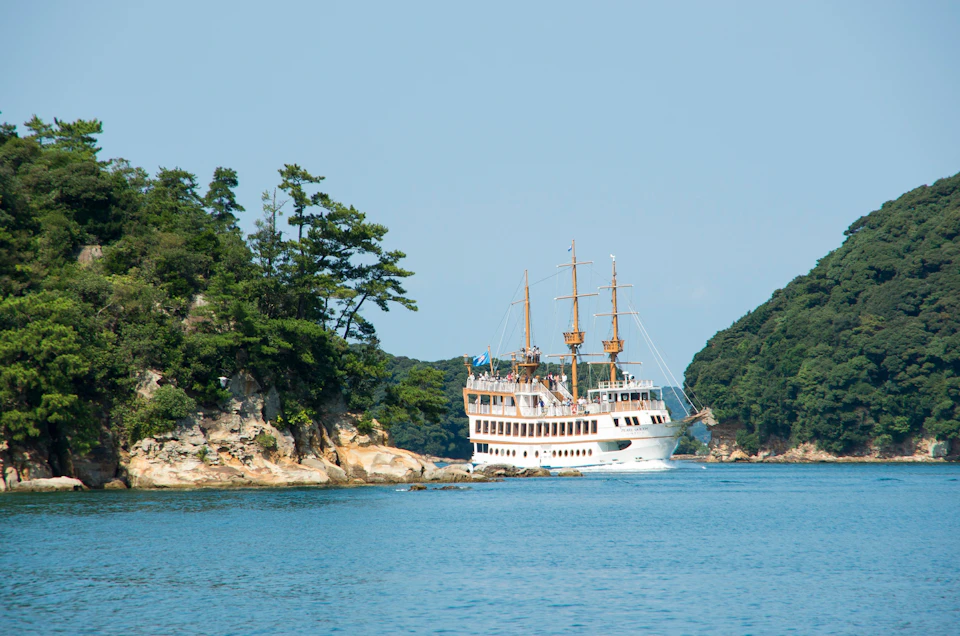 A white ship with sails navigates near a forested coastline under a clear blue sky.