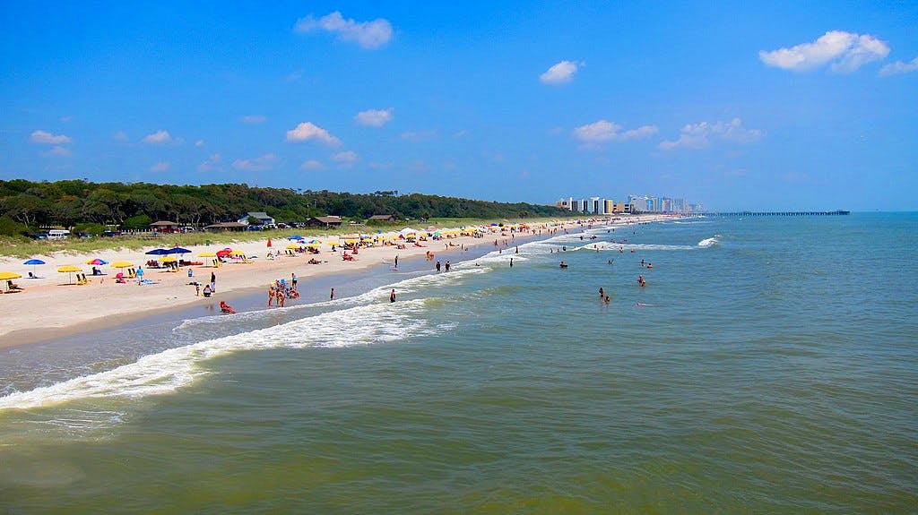 Beach scene with people sunbathing and swimming. Colorful umbrellas dot the sandy shore, buildings in the distance, under a blue sky.