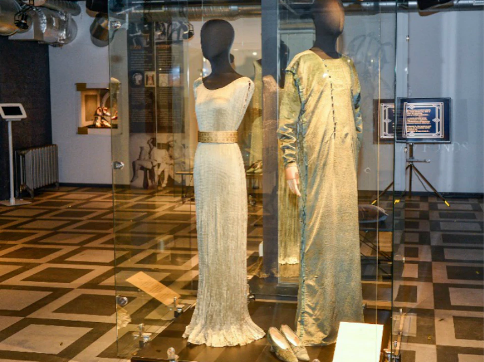 Two mannequins in glass cases display a white evening gown and a long-sleeved green dress with matching shoes.