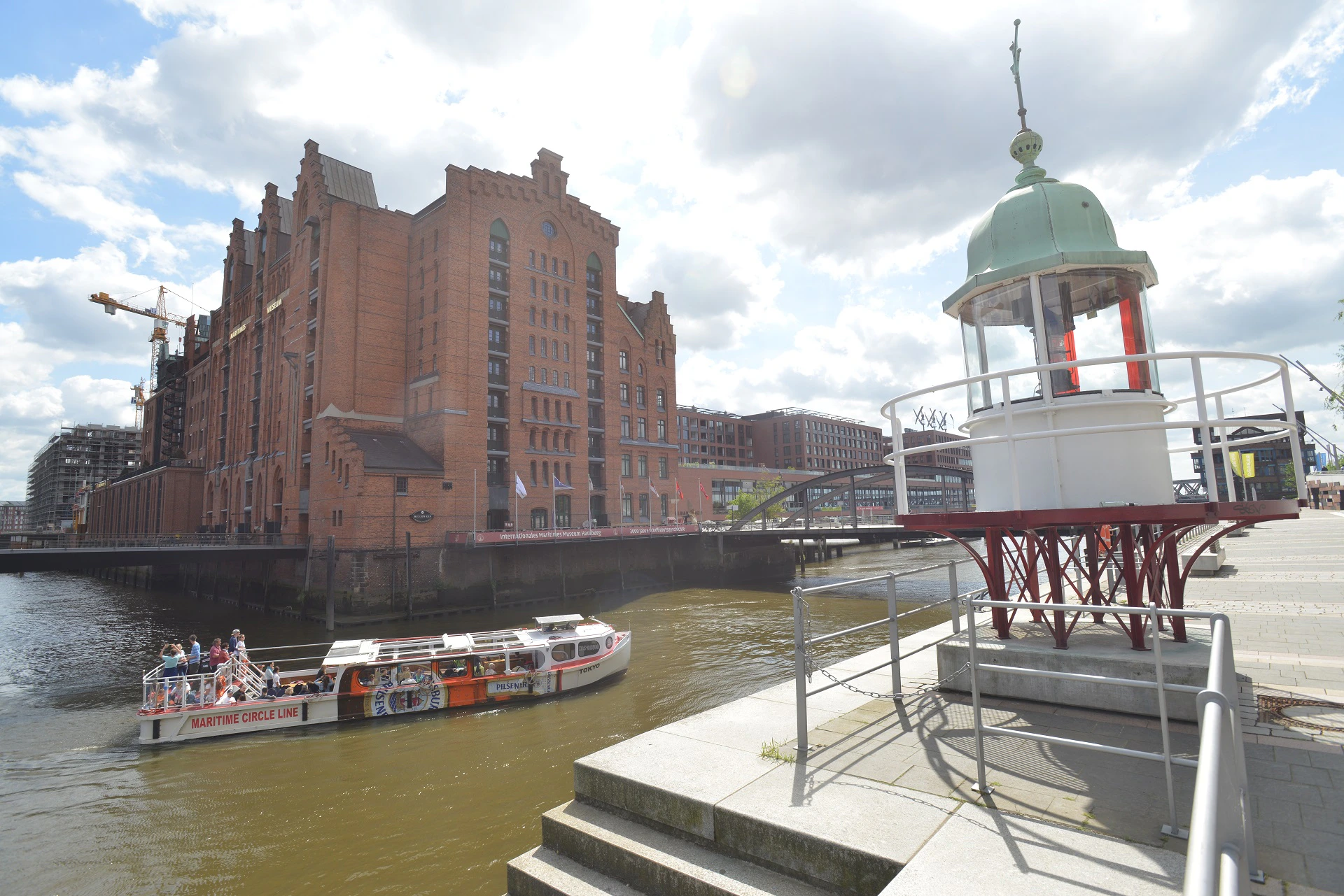 Brick waterfront buildings, a boat floating in the canal, and a white tower with a green dome in the foreground under a cloudy sky.