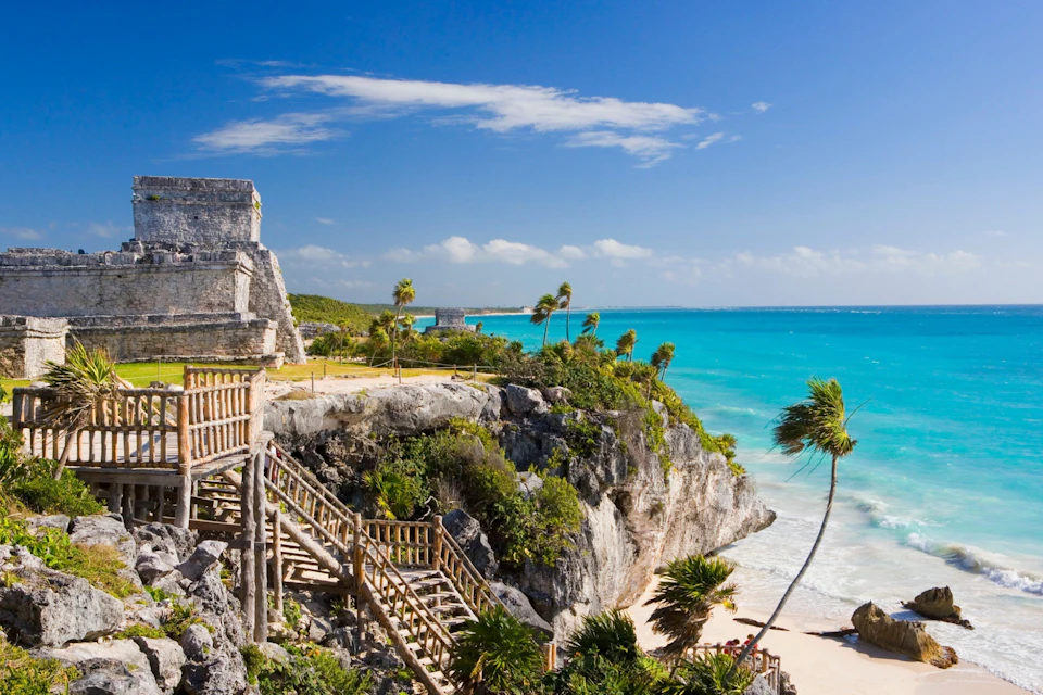 Stone ruins and wooden stairs on a cliff overlooking a turquoise beach, with palm trees and a partly cloudy blue sky.
