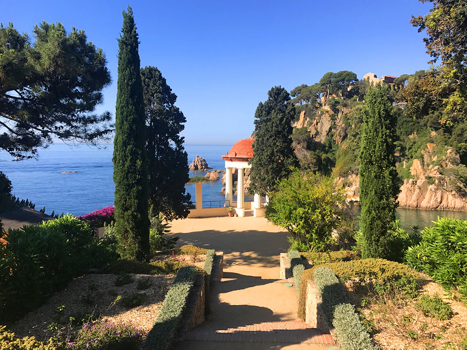 A landscaped garden overlooking the sea with a red-roofed pavilion, tall trees, and rocky cliffs under a clear blue sky.