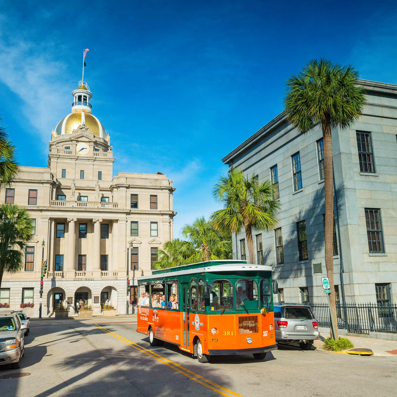 A green and orange trolley drives past a building with a gold dome under a clear blue sky, with palm trees lining the street.