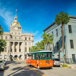 A green and orange trolley drives past a building with a gold dome under a clear blue sky, with palm trees lining the street.