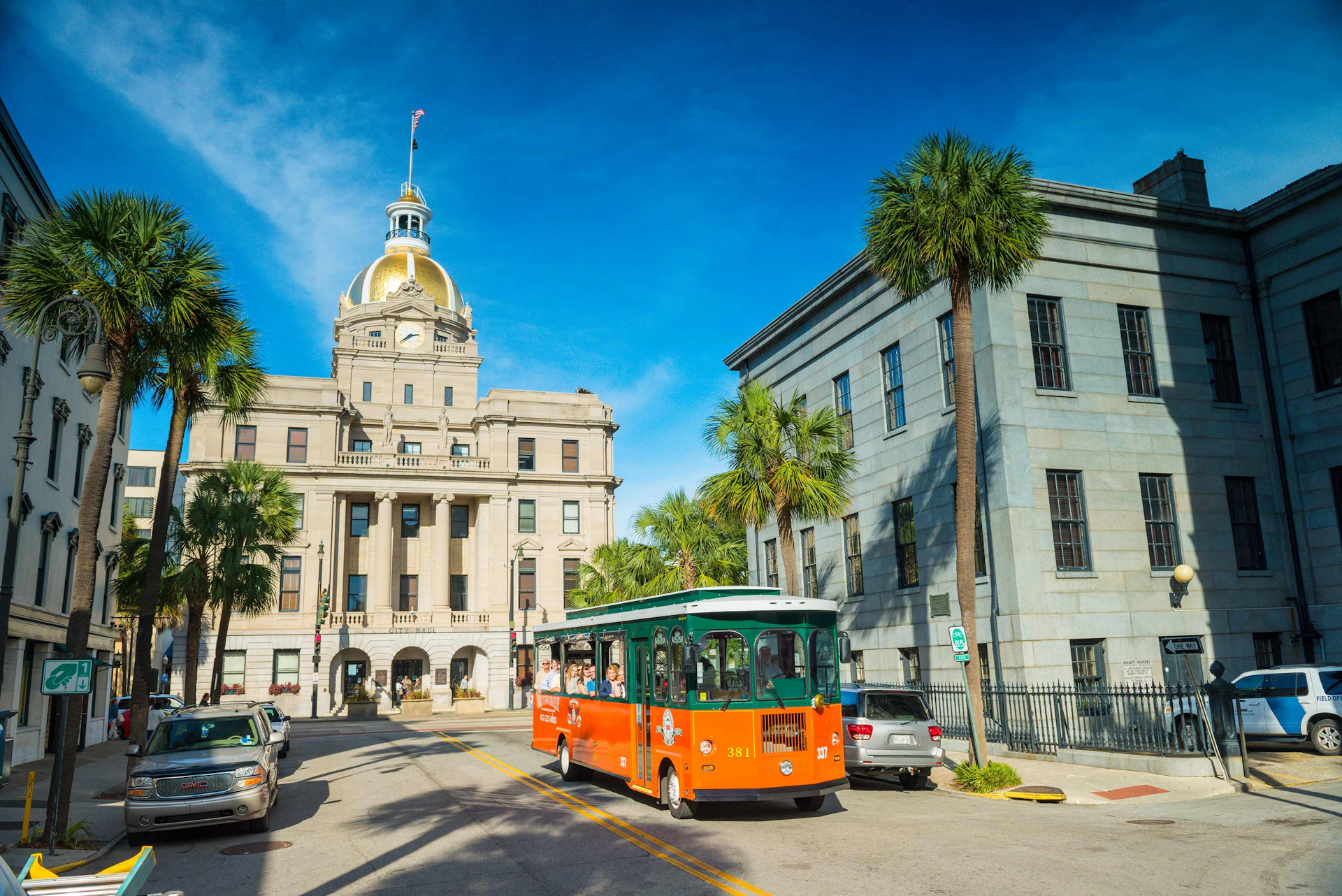 A green and orange trolley drives past a building with a gold dome under a clear blue sky, with palm trees lining the street.