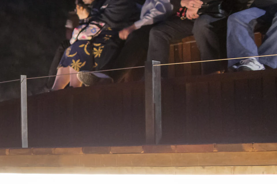 People observing penguins at a beach from an elevated seating area and a glass-walled viewing area at dusk.