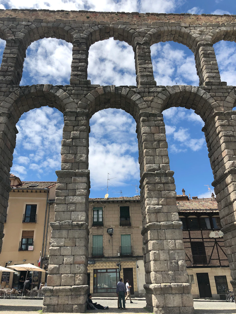 Ancient stone archway with multiple arches in front of old buildings under a blue sky with scattered clouds.