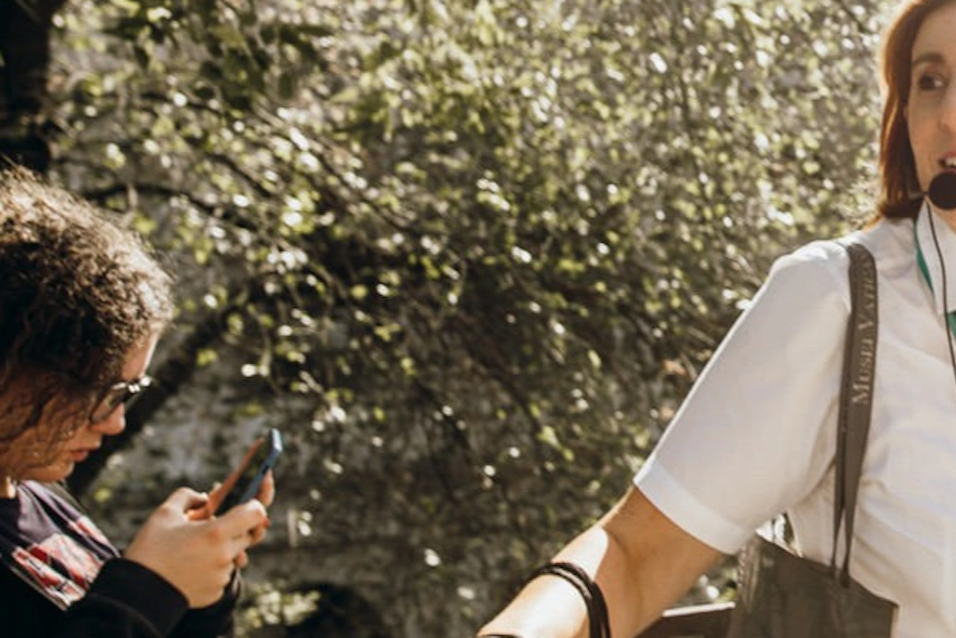 A woman wearing a microphone headset leads a tour near an ancient structure, with trees and sunlight in the background.