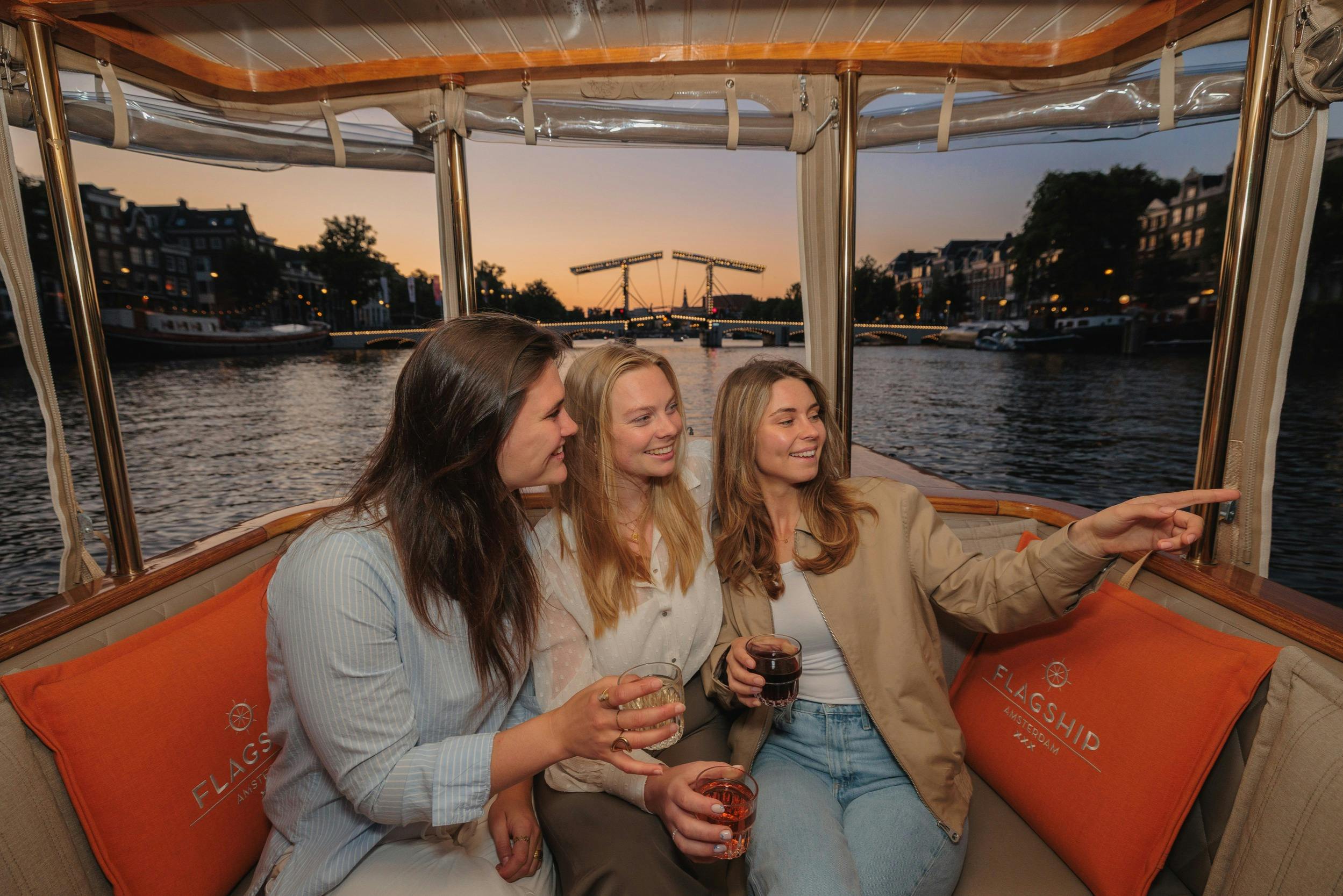 Three women on a boat enjoying drinks at sunset, with one pointing toward a bridge over a river.