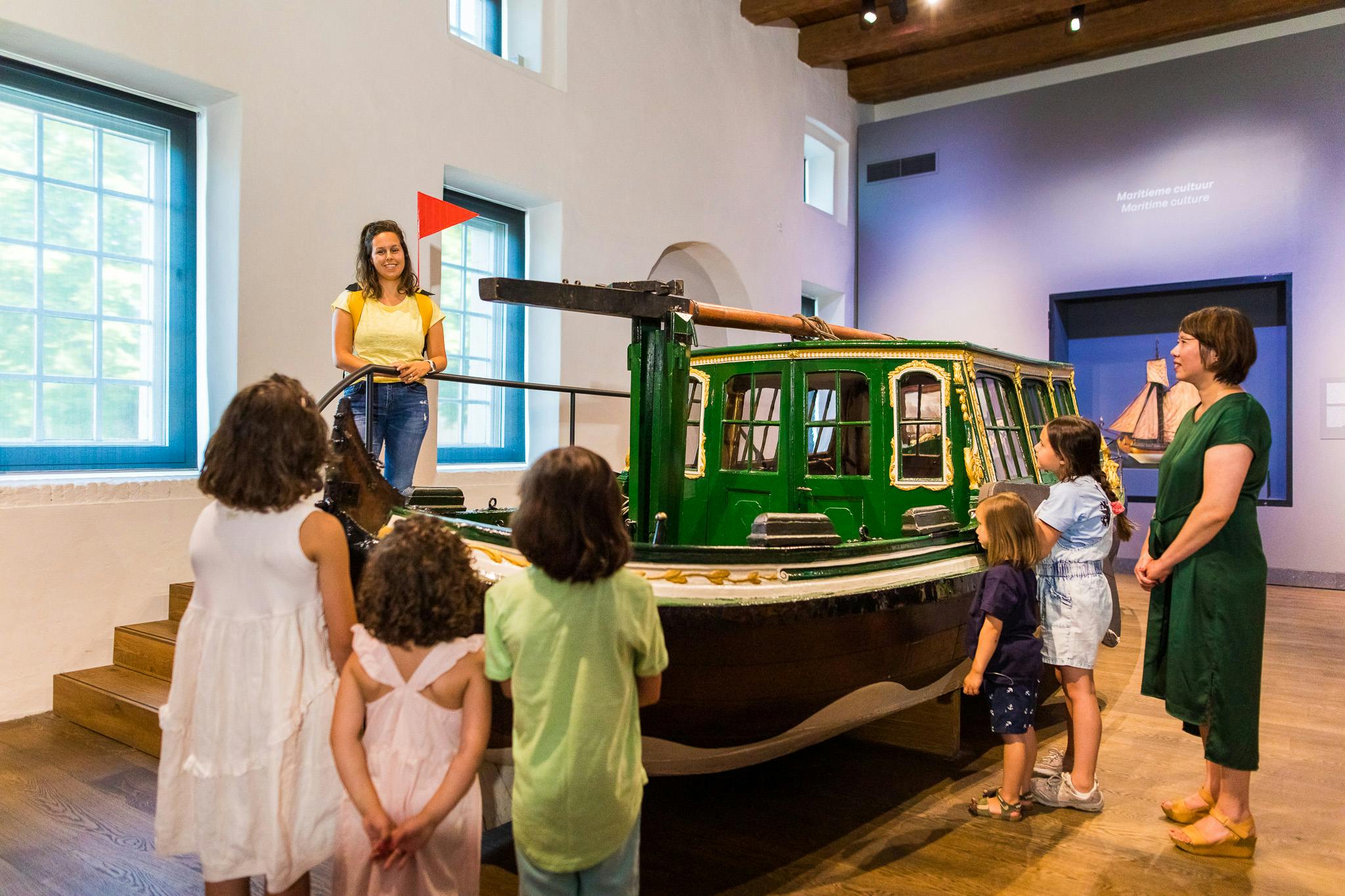 Una donna si trova vicino a una barca verde all'interno di una stanza mentre tre bambini guardano. La stanza ha grandi finestre e travi in legno.