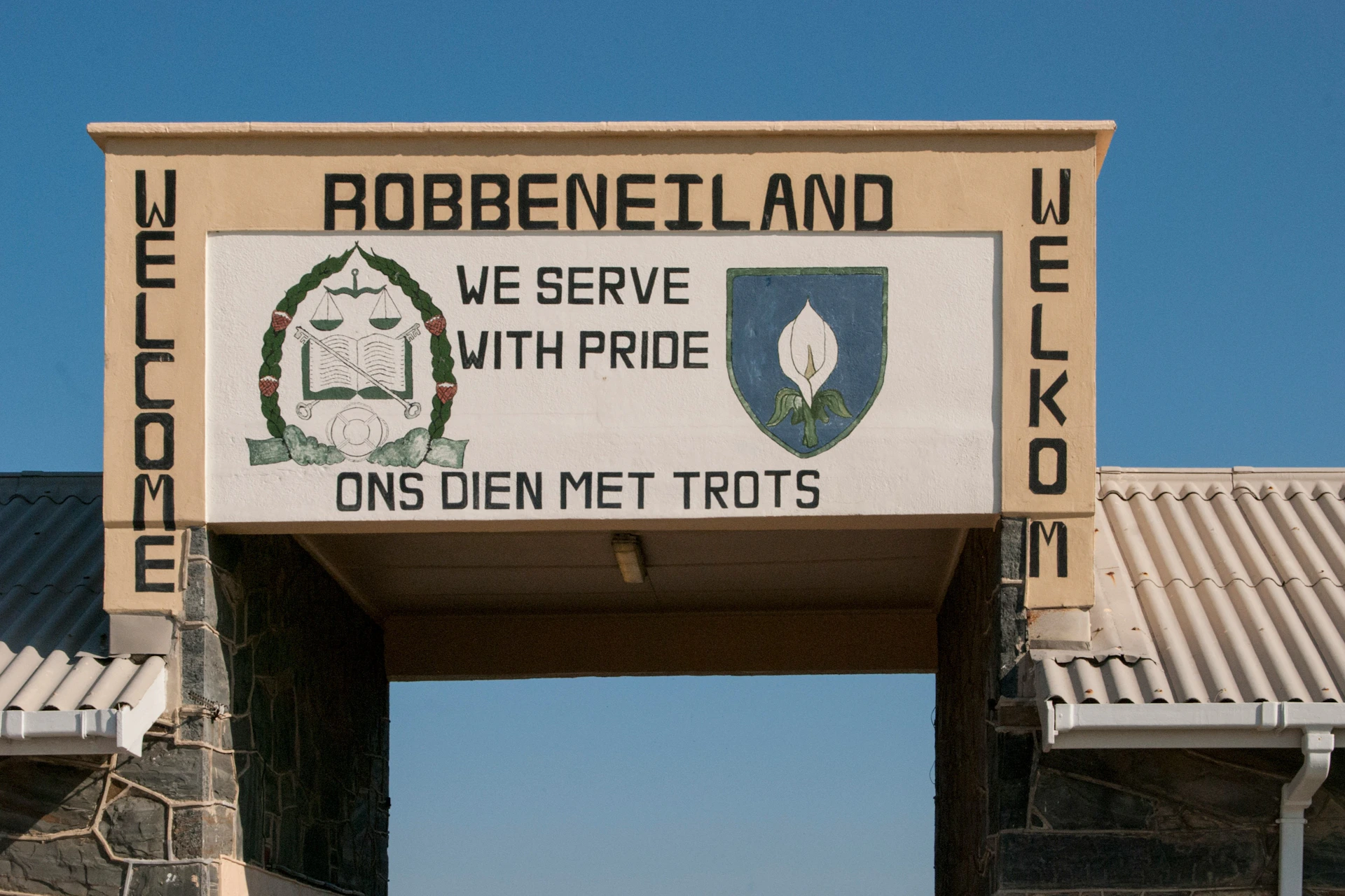 An entrance sign at Robben Island with the text "WE SERVE WITH PRIDE / ONS DIEN MET TROTS" and "WELKOM" on the side.