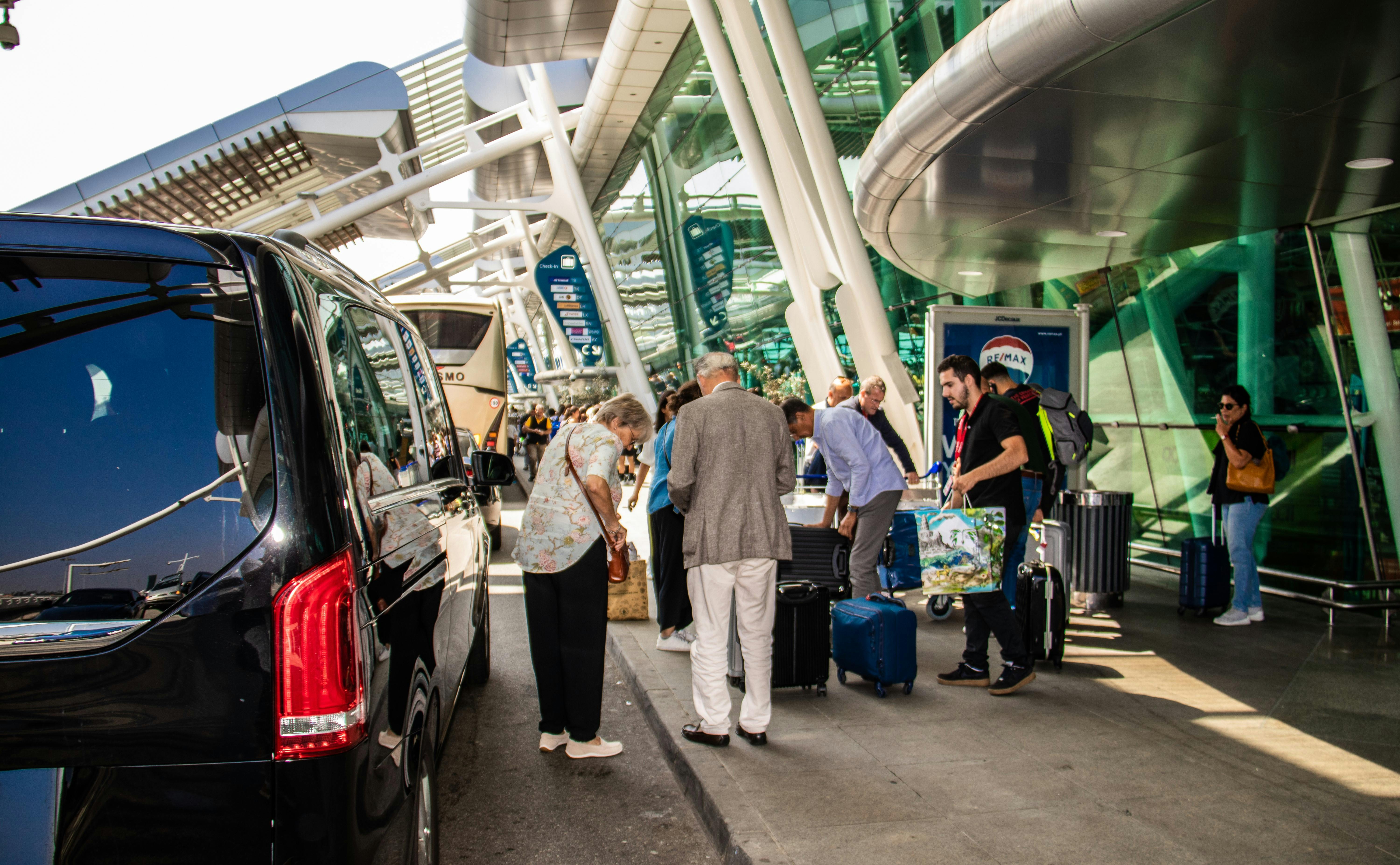 Travelers with luggage standing near vehicles outside a modern glass airport terminal building.