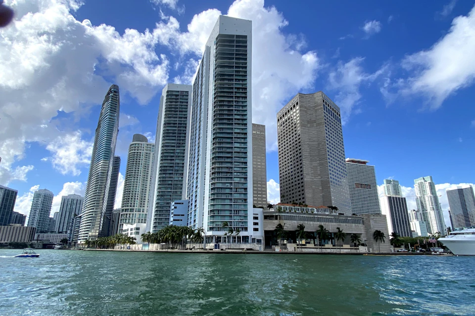 Tall modern skyscrapers line a waterfront under a partly cloudy blue sky, with palm trees and water in the foreground.