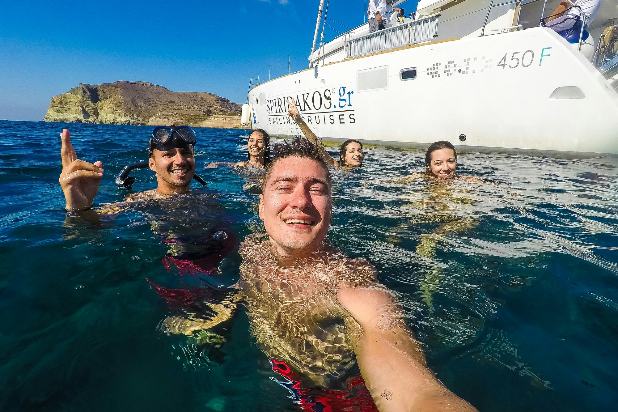 맑고 푸른 하늘 아래 측면에 "세일링 크루즈" 라고 쓰여진 하얀 보트 근처에서 5명이 수영하고 미소를 짓고 있습니다.