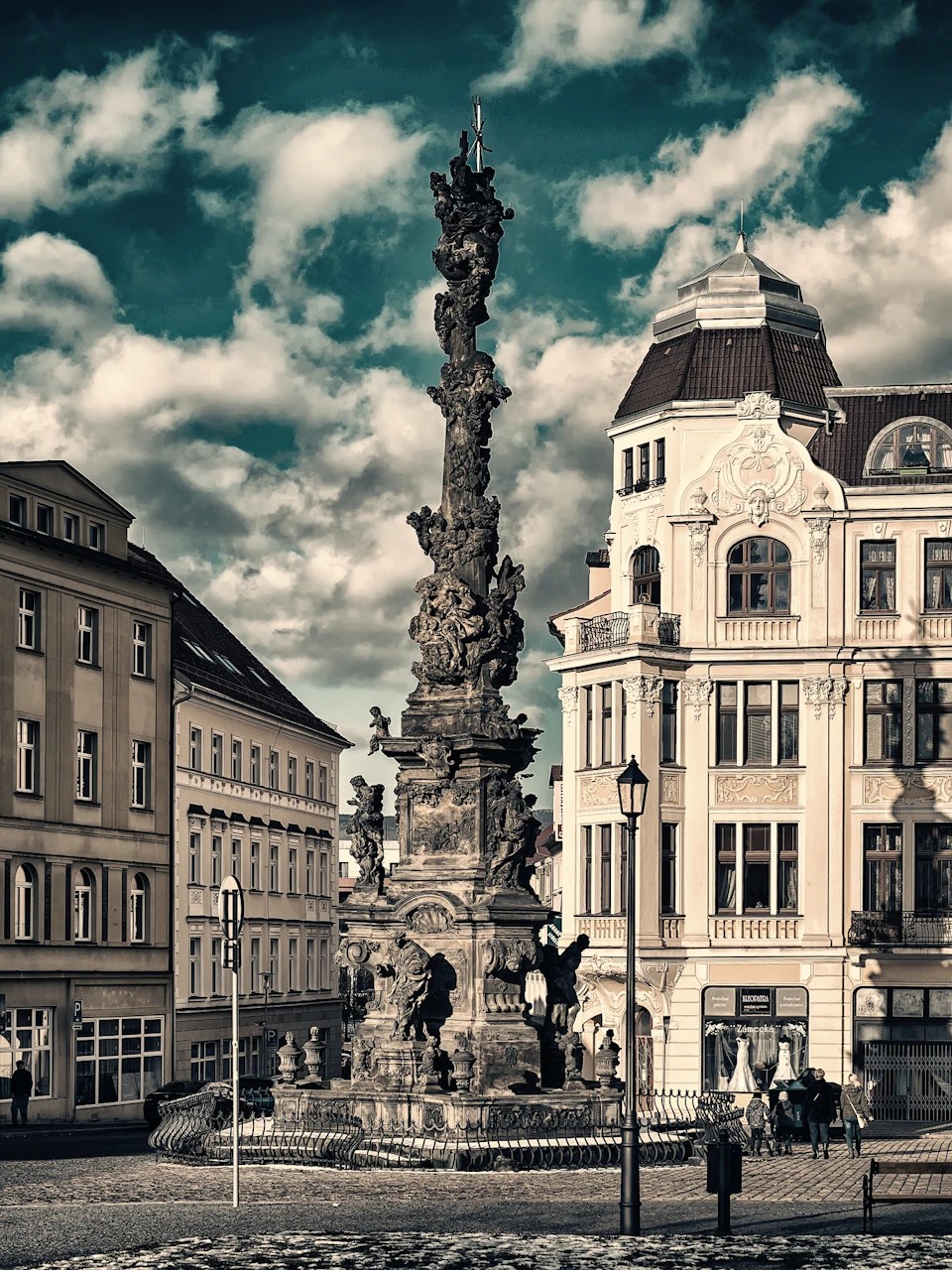 Tall, intricate monument with statues in front of ornate buildings under a cloudy sky.