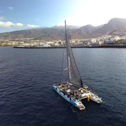 A catamaran with a single black sail on calm waters, with a coastal town and mountain range in the background under a blue sky.