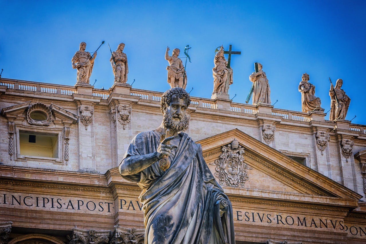 A large statue in front of a historic building with Latin inscriptions and four statues on the roof under a clear blue sky.