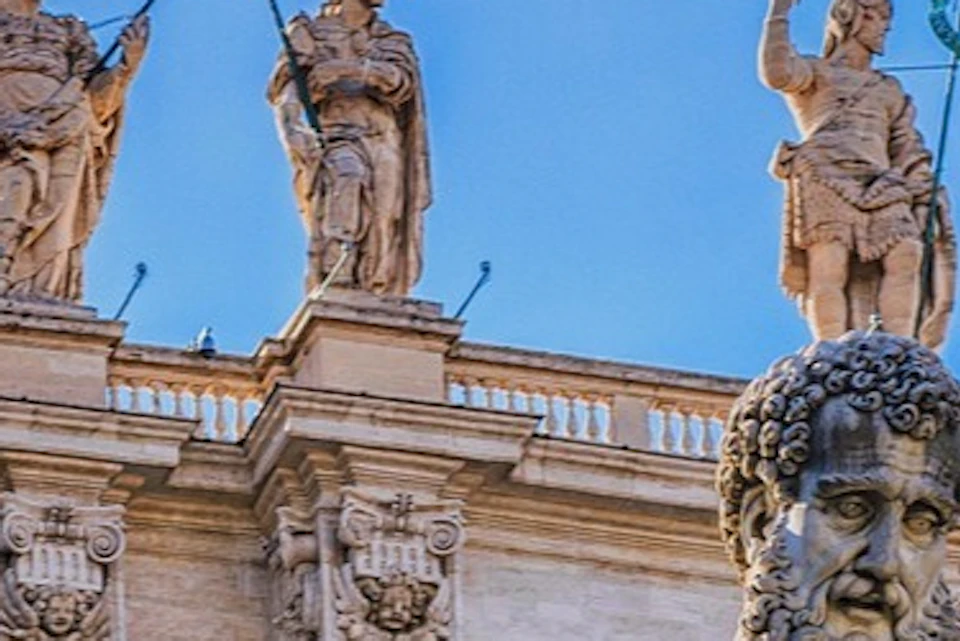 A large statue in front of a historic building with Latin inscriptions and four statues on the roof under a clear blue sky.