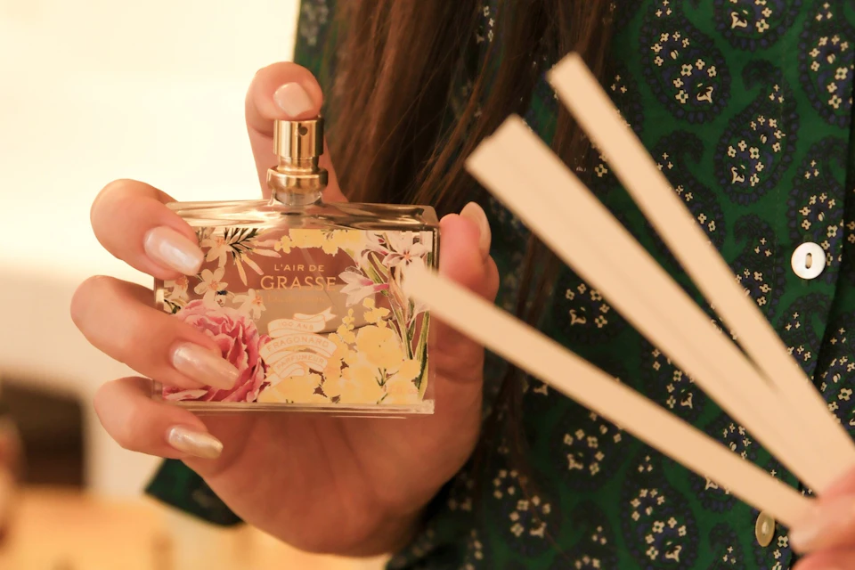A person holding a floral-printed perfume bottle labeled 'L'air de Grasse,' with wooden scent sticks in the foreground.