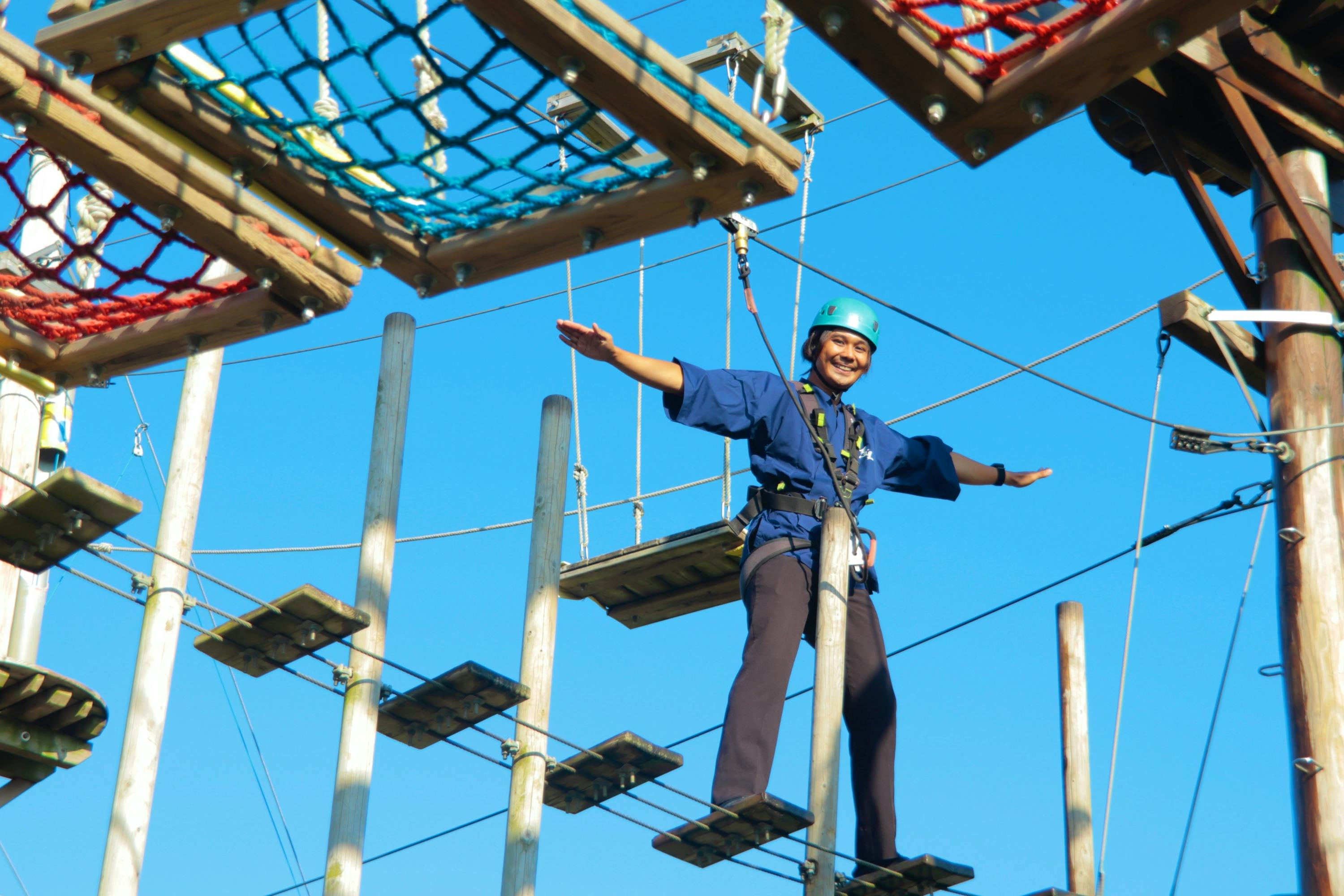 A person wearing safety gear is balancing on a high ropes course under a clear blue sky.