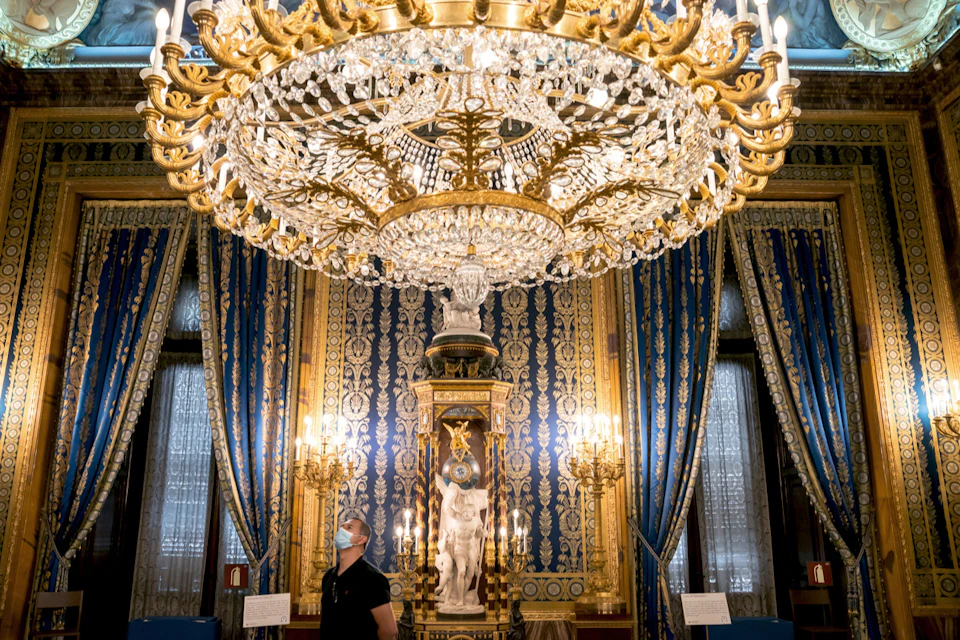 A lavish room with a large ornate chandelier, blue and gold decor, a statue on a pedestal, and a person wearing a mask.