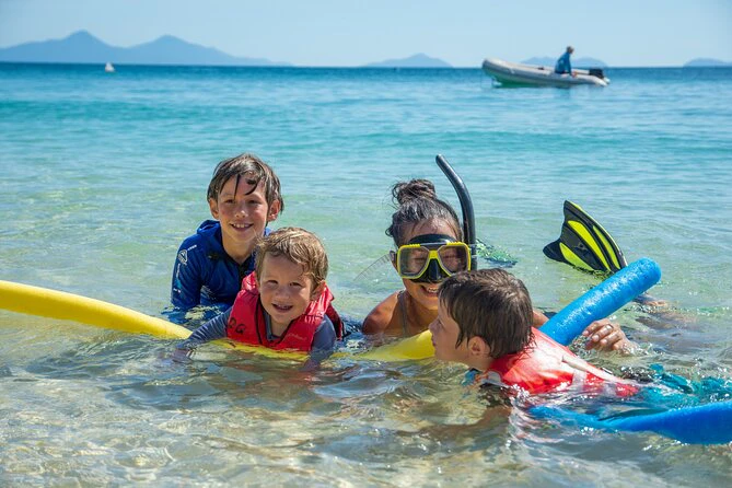 Four children in life vests playing in shallow ocean water, one wearing a snorkel mask. Mountains and a boat are in the background.