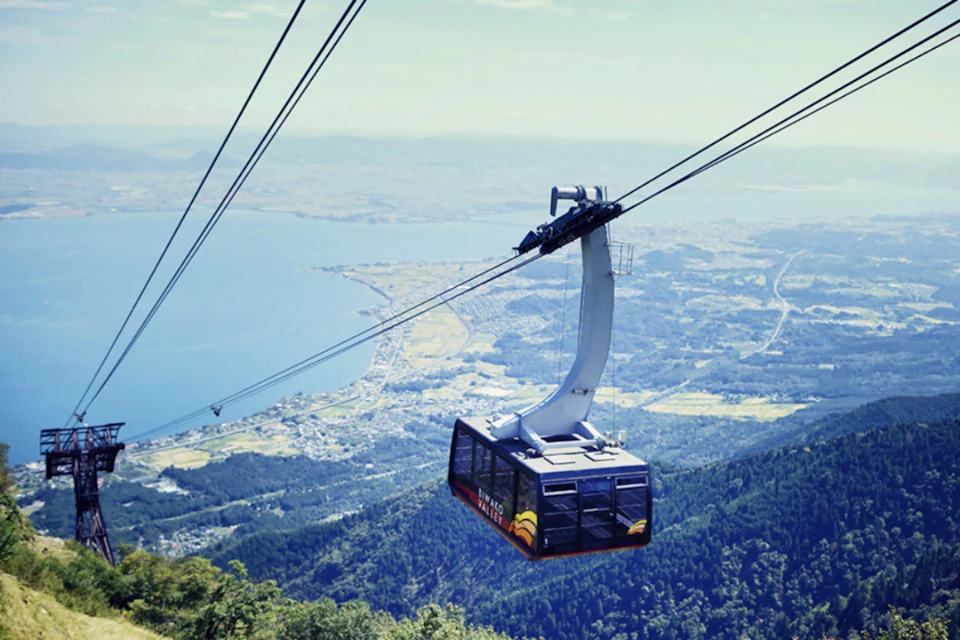 A cable car traveling over a forested mountain with a view of a lake and a distant townscape under a clear sky.