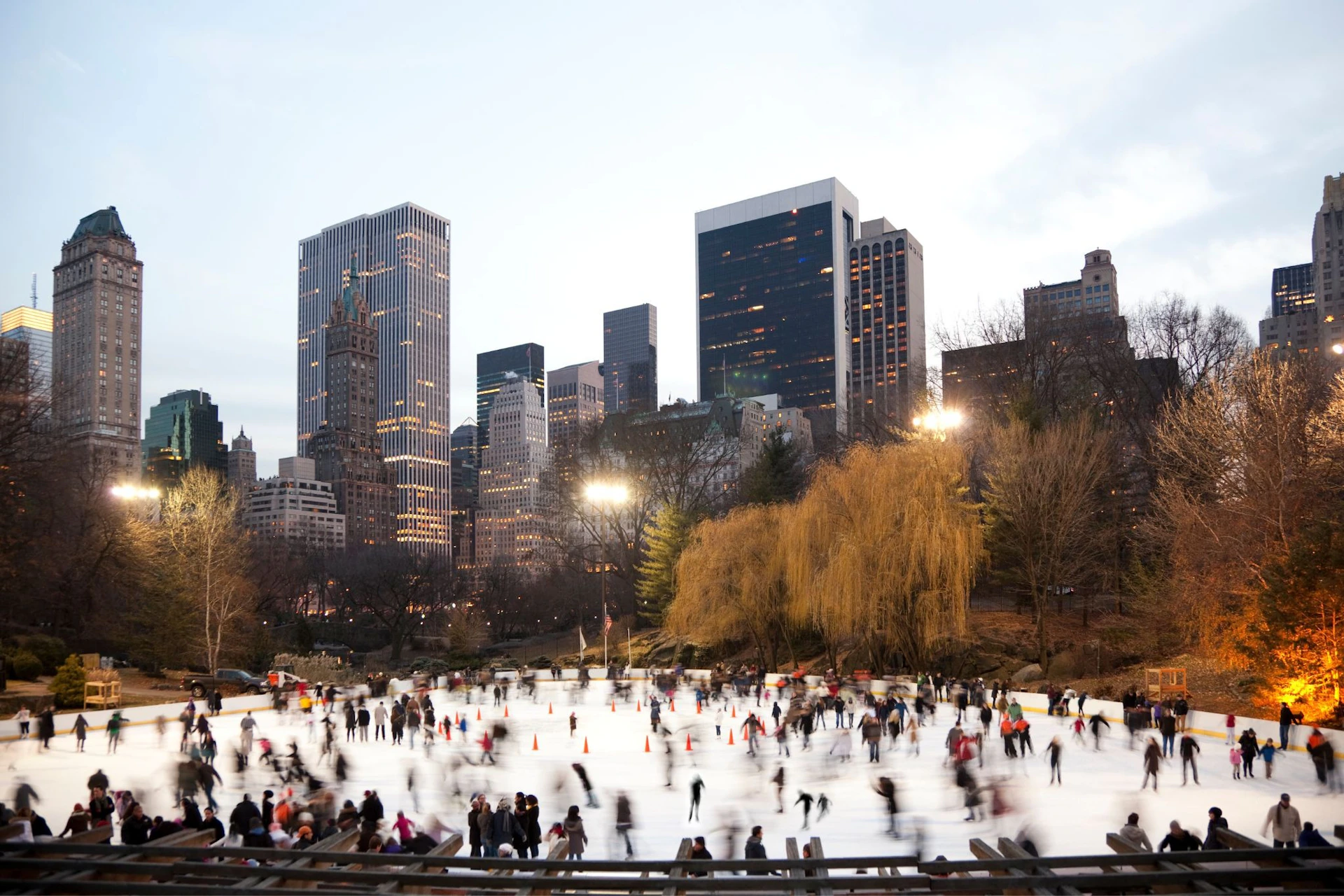 People ice skating in a park surrounded by tall buildings and trees. The scene is lit by floodlights with a city skyline in the background.