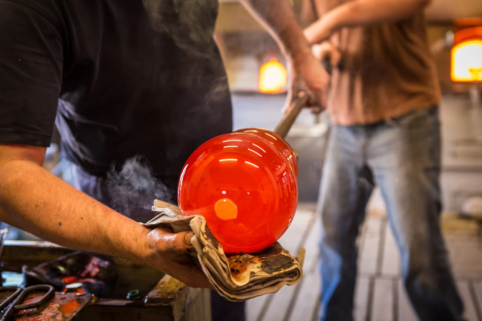 A person shapes a glowing, hot glass object using a rod and newspaper while another person works in the background.