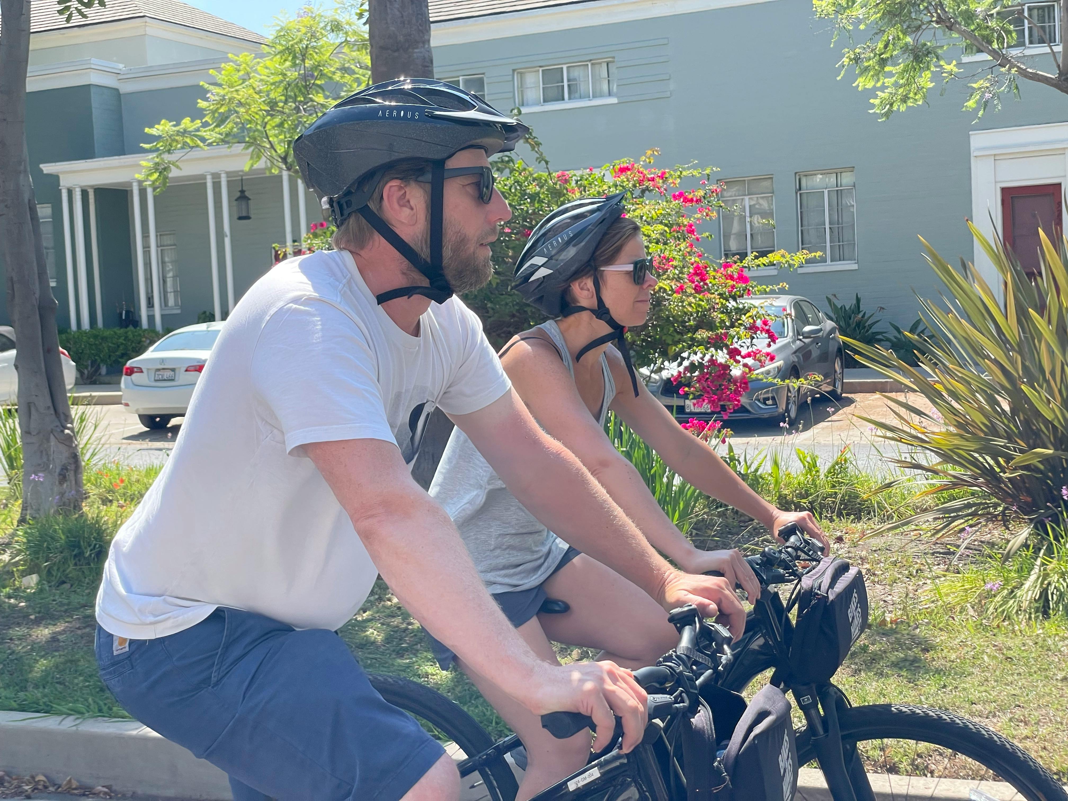 Two people wearing helmets and sunglasses ride bicycles on a sunny street, with a house and flowering bushes in the background.