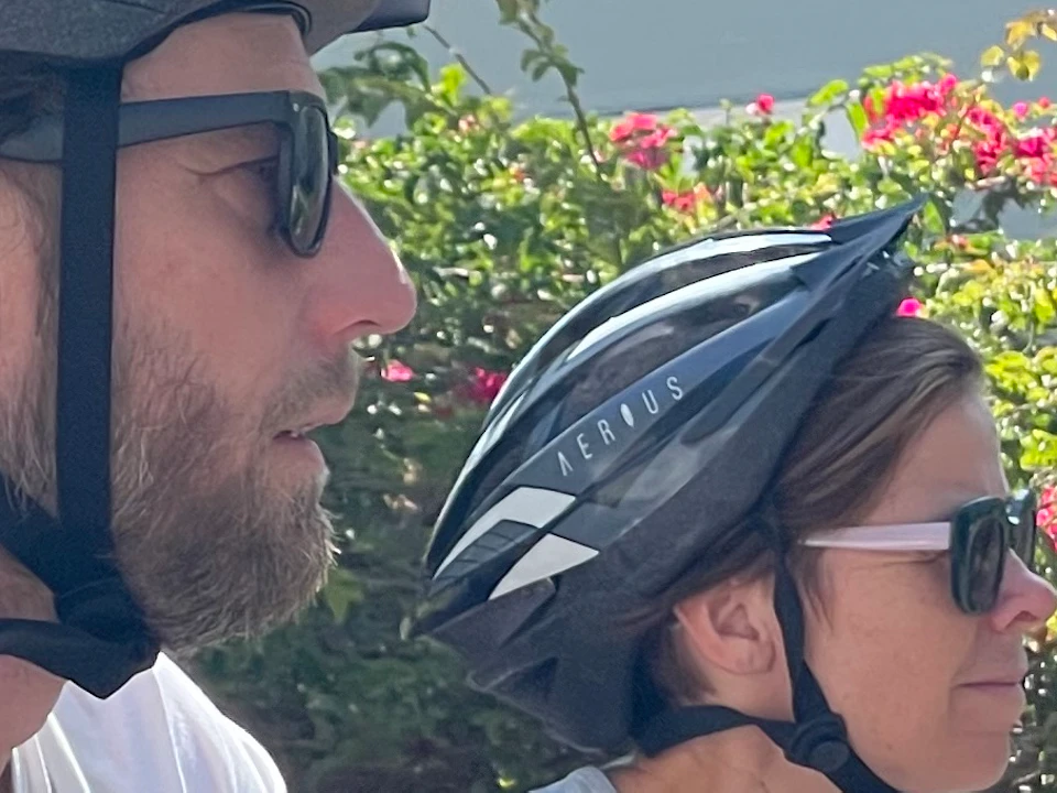 Two people wearing helmets and sunglasses ride bicycles on a sunny street, with a house and flowering bushes in the background.