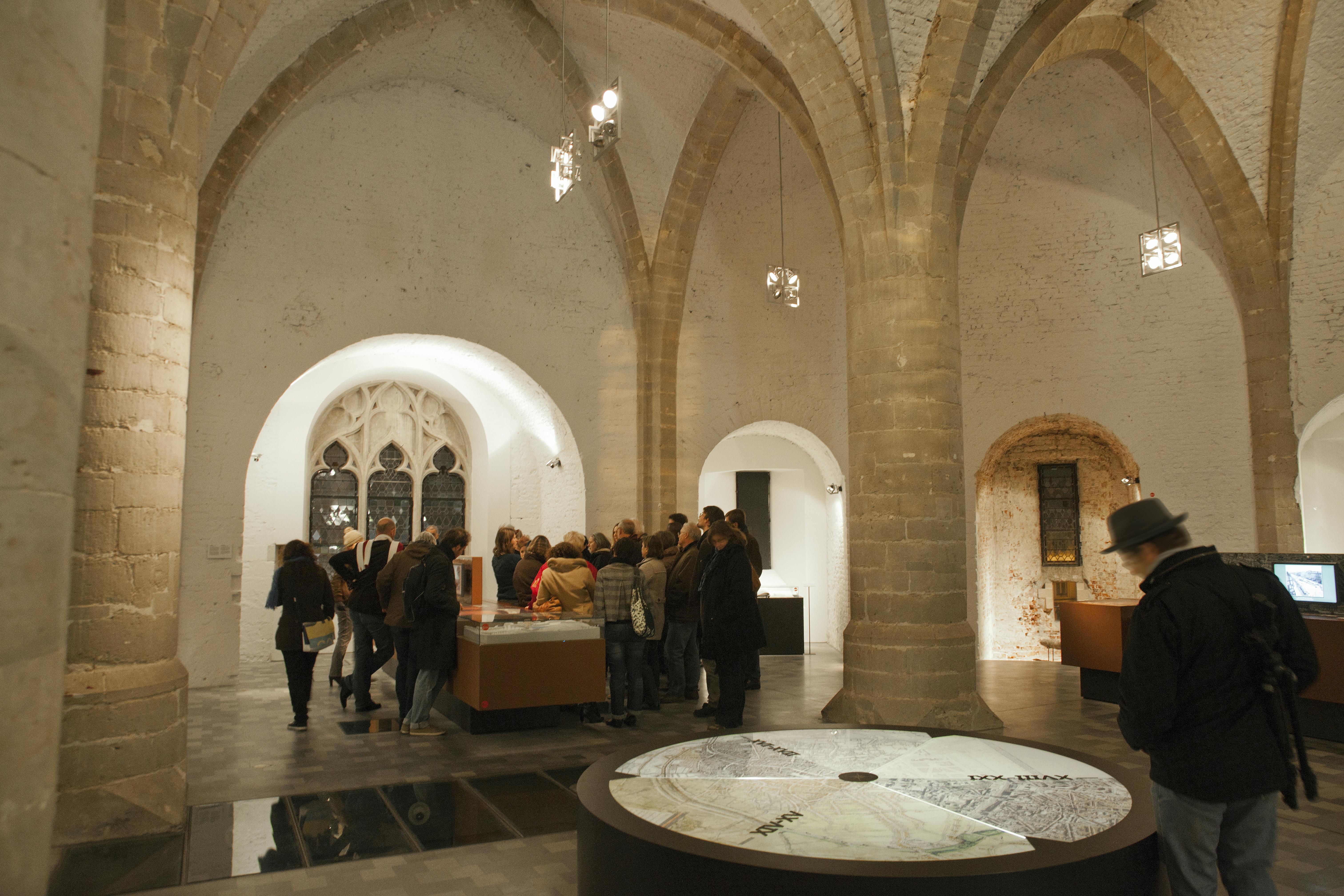游客们聚集在一个具有拱形天花板和石柱的历史悠久的博物馆的展品周围。 前景中有一张圆形展示桌。