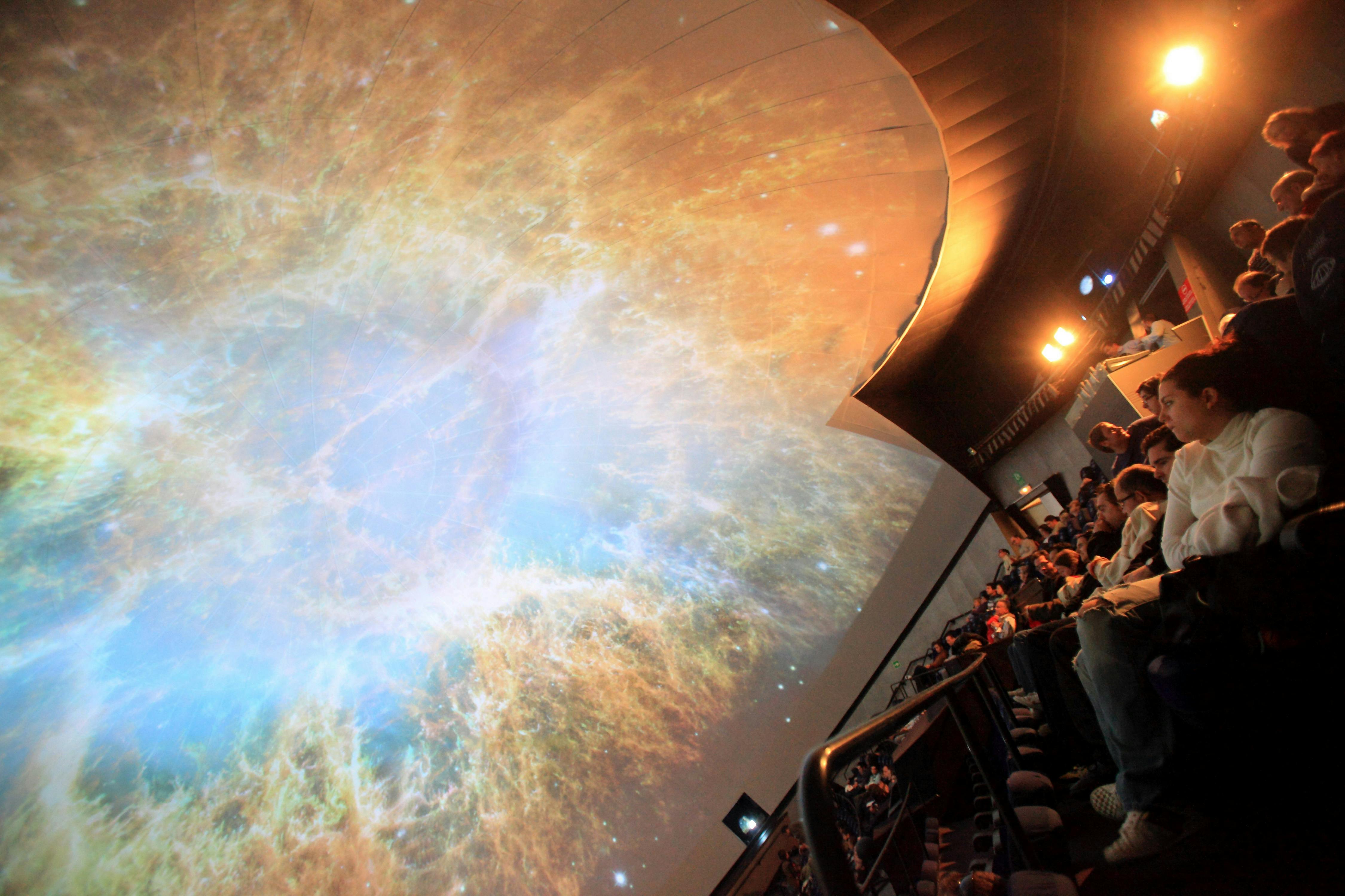 Lidé sedí v planetáriu a sledují velkou projekci barevné mlhoviny na klenutém stropě.