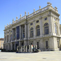 En storslagen, utsmyckad byggnad med pelare, statyer på taket, färgglada flaggor och en staty i förgrunden under en klarblå himmel.