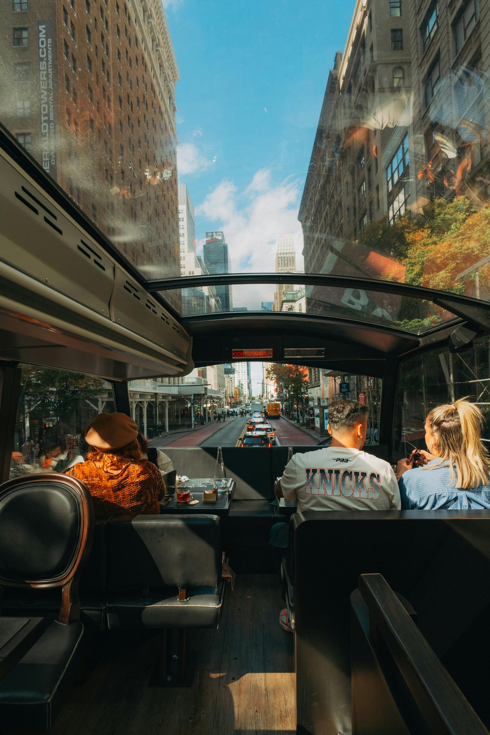 Mensen zitten in een bus met een glazen dak en kijken uit op een stadsstraat met hoge gebouwen, verkeer en een blauwe lucht.