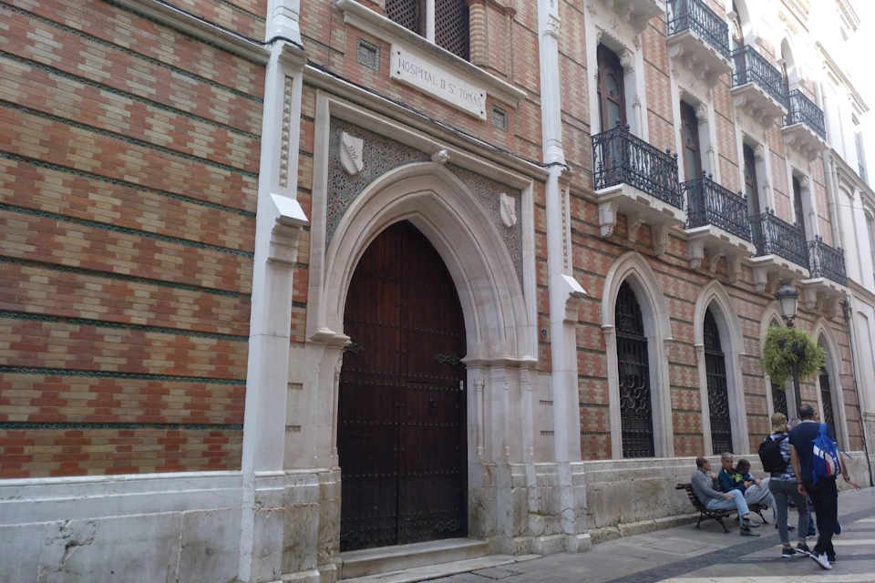 Ornate brick building with arched wooden door labeled "Hospital de Todos." Iron balconies and windows are visible.