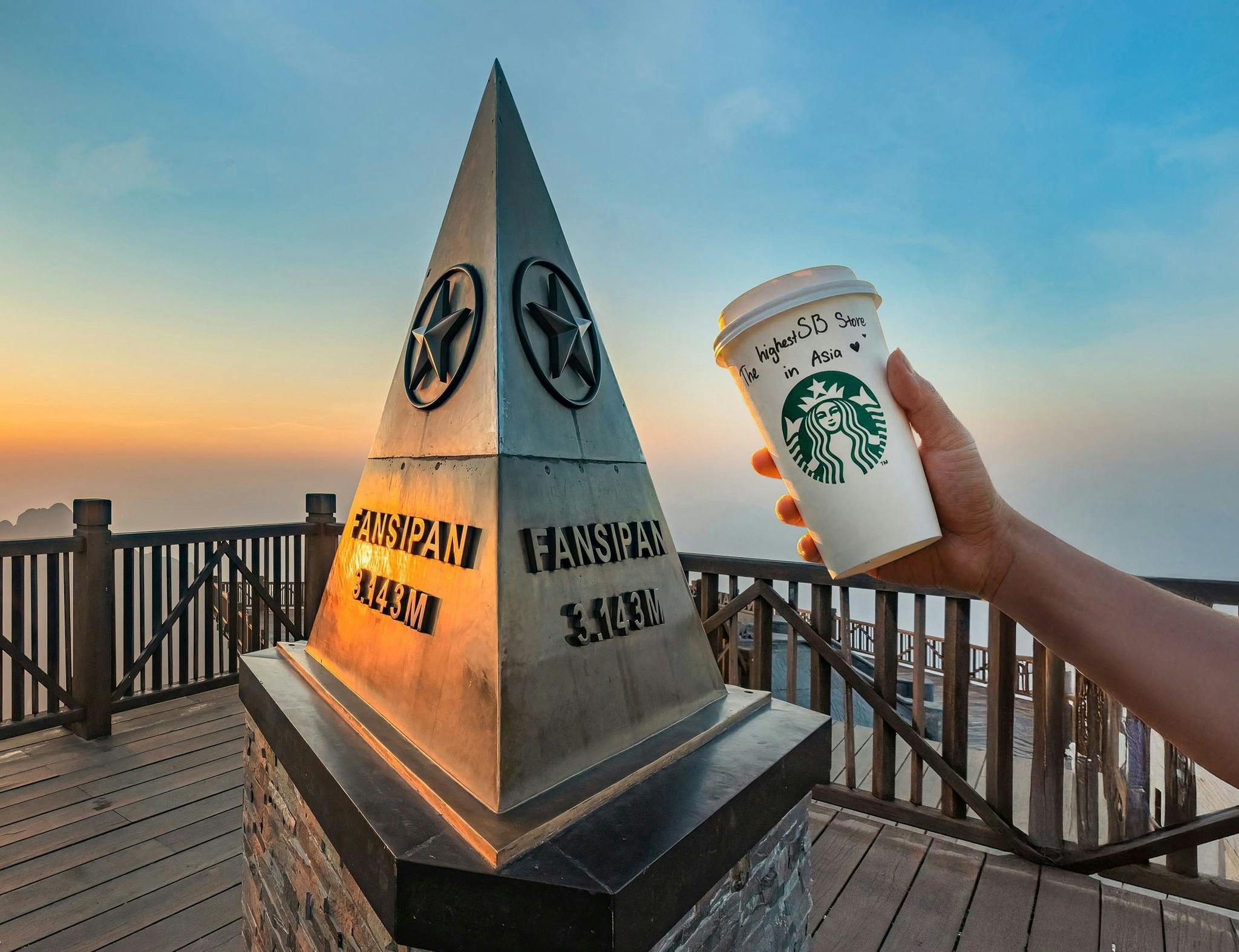A hand holding a Starbucks cup near a triangular metal monument on a mountain, labeled Fansipan 3,143m, at sunrise.