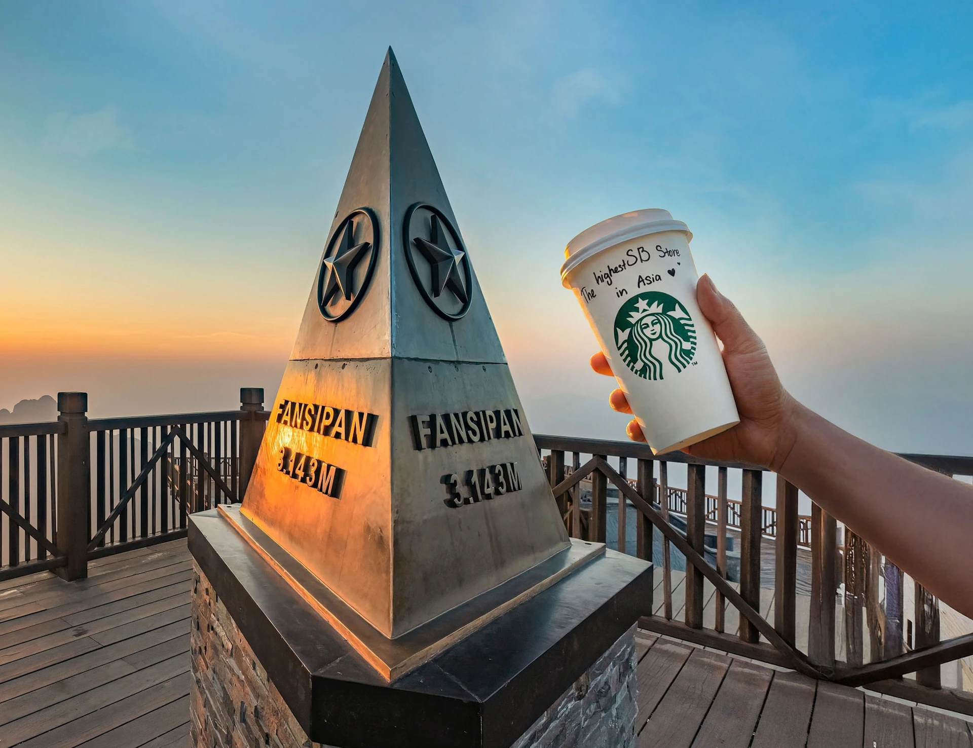 A hand holding a Starbucks cup next to a triangular Fansipan summit marker at sunset, with a scenic mountain view.