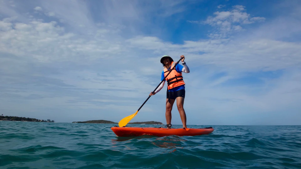 Person standing on an orange paddleboard in the ocean, holding a yellow paddle, wearing a life jacket and hat, with a blue sky backdrop.