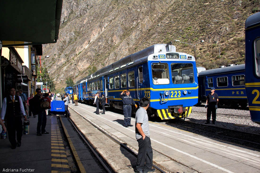Το μπλε και άσπρο τρένο με τον αριθμό 223 βρίσκεται σε έναν σταθμό δίπλα στα βουνά. Αρκετοί άνθρωποι στέκονται δίπλα στο τρένο.