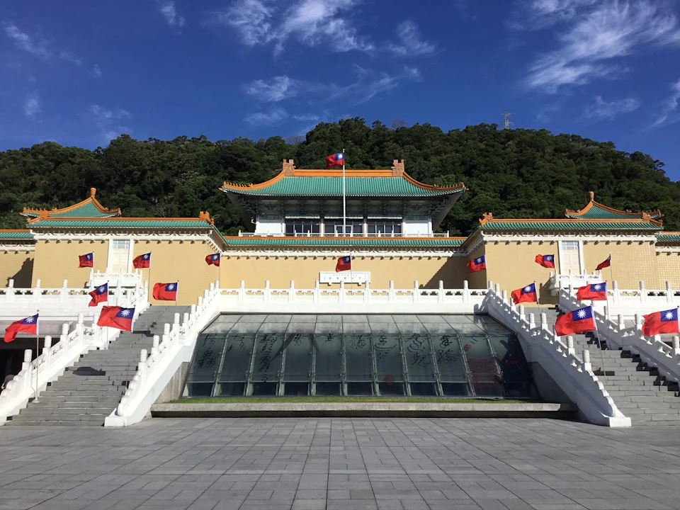 A traditional Chinese building with green rooftops, surrounded by national flags, sits against a backdrop of trees under a blue sky.