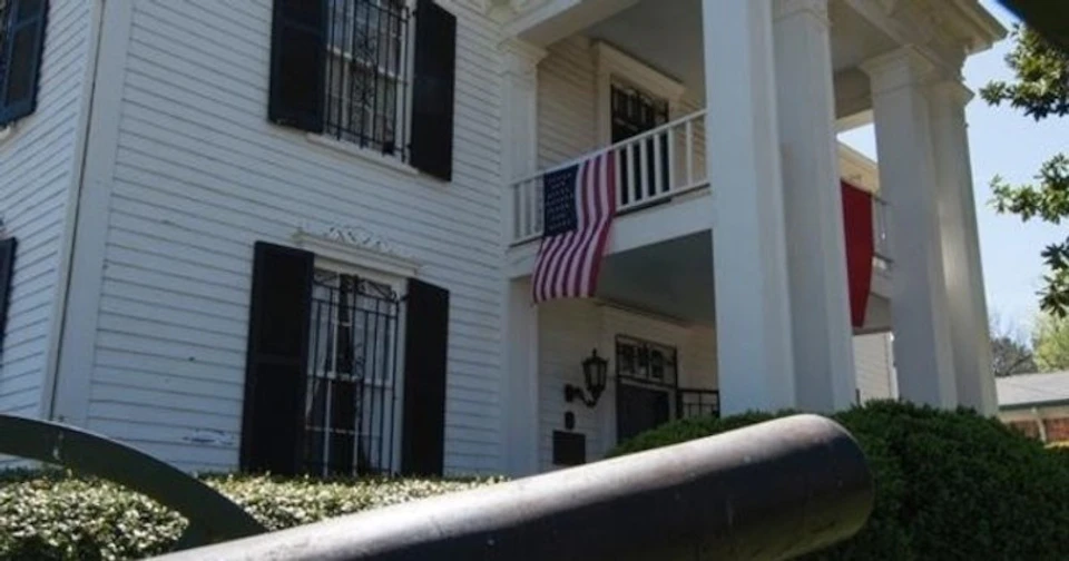 White building with black shutters, a United States flag hanging from a balcony, and ornamental railings on windows.