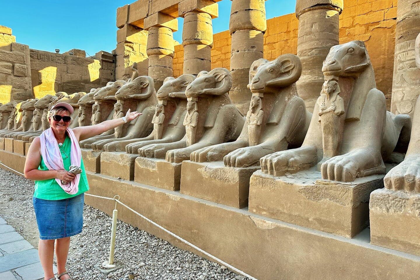 A woman poses, pointing at a row of ram-headed sphinx statues in an ancient stone temple under a clear blue sky.