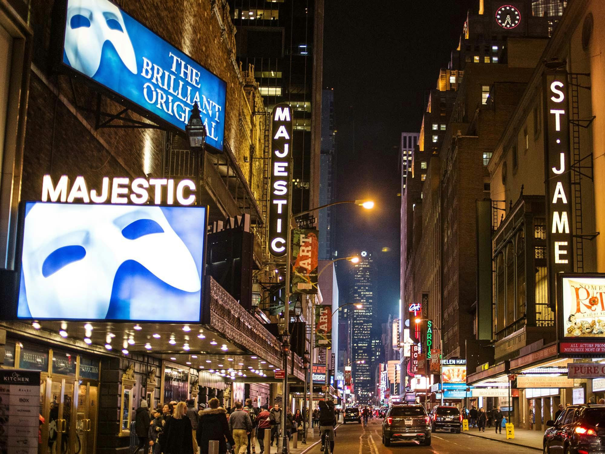 Busy nighttime street with theater marquees and billboards displaying "Majestic" and "The Brilliant Original." Pedestrians and cars present.