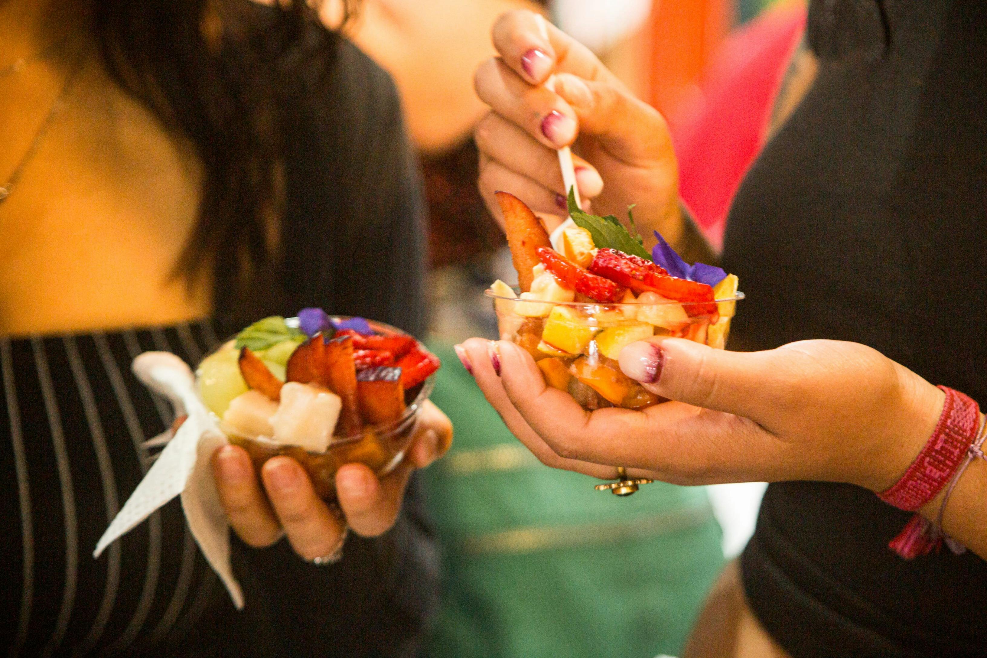 Zwei Personen halten Schüsseln mit buntem Fruchtsalat mit Erdbeeren, Pflaumen und essbaren Blumen, eine davon mit einem Löffel zum Essen.