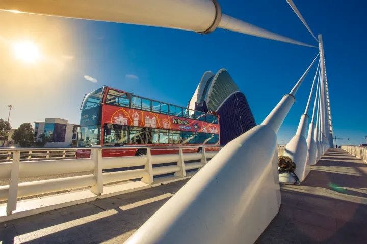 Κόκκινο διώροφο λεωφορείο σε μια σύγχρονη λευκή γέφυρα με ένα μοναδικό αρχιτεκτονικό κτίριο στο βάθος κάτω από έναν καταγάλανο ουρανό.