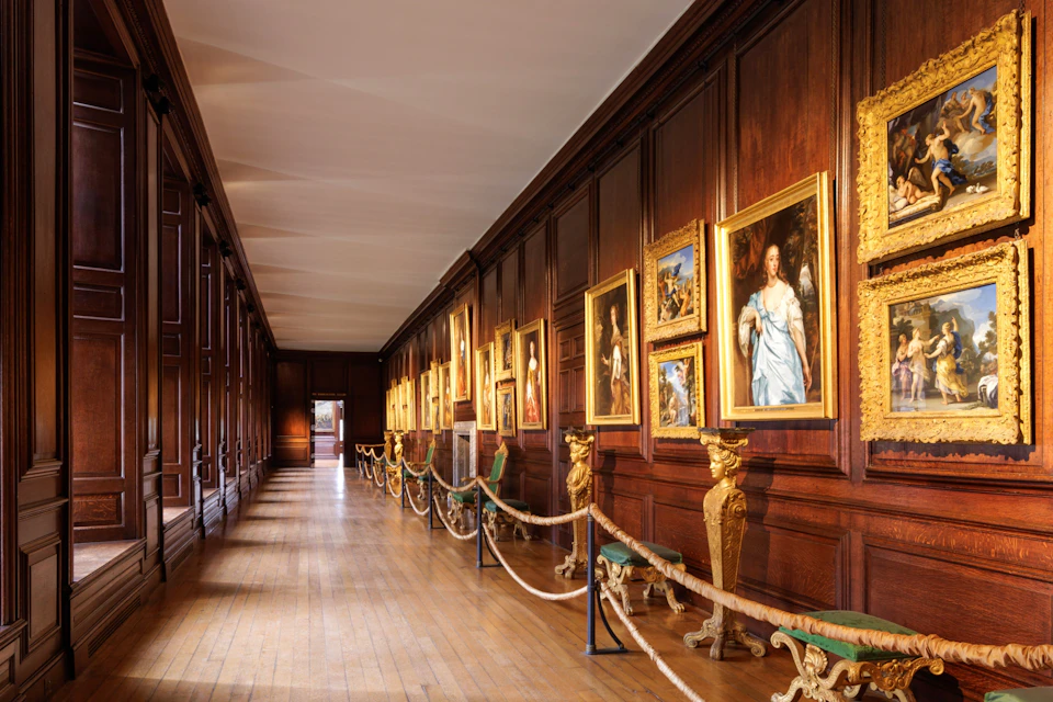 A long gallery with wooden paneling featuring framed paintings, ornate benches, and a roped-off walkway.